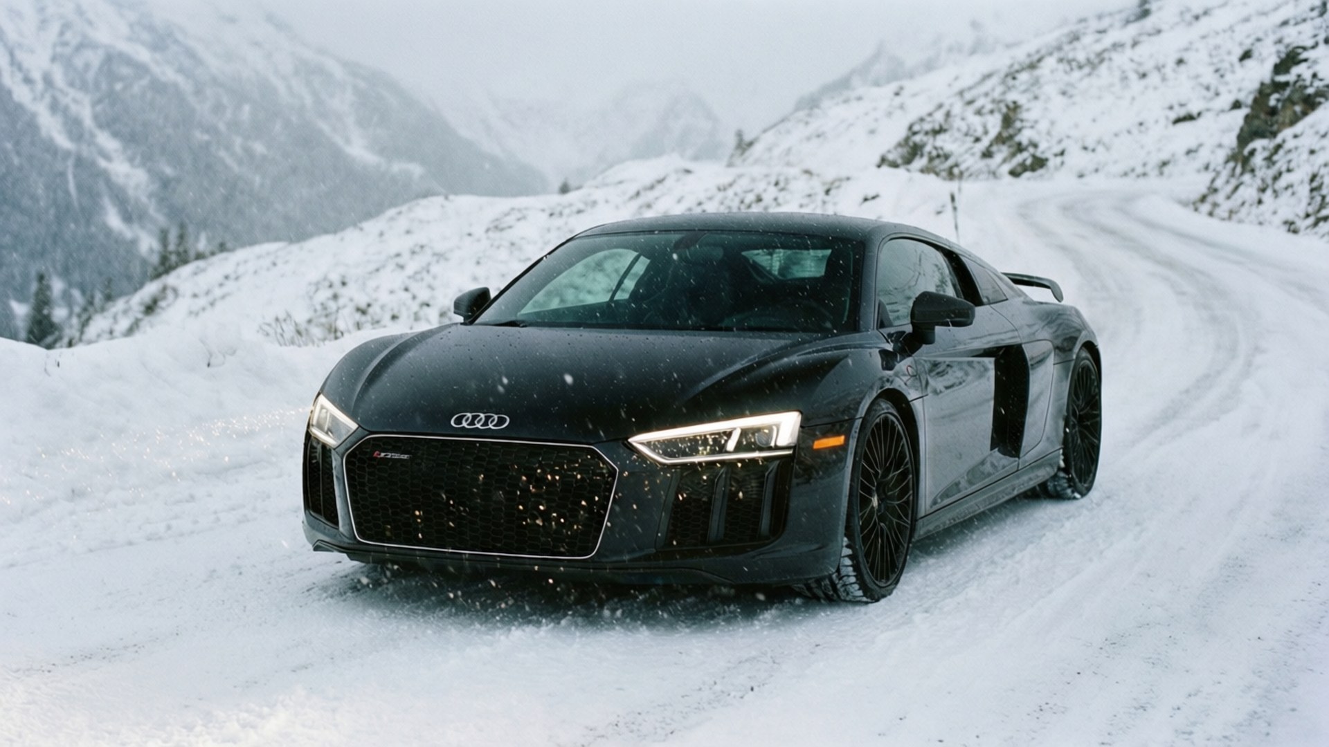Черно Audi R8 се движи по заснежен алпийски път сред падащ сняг.